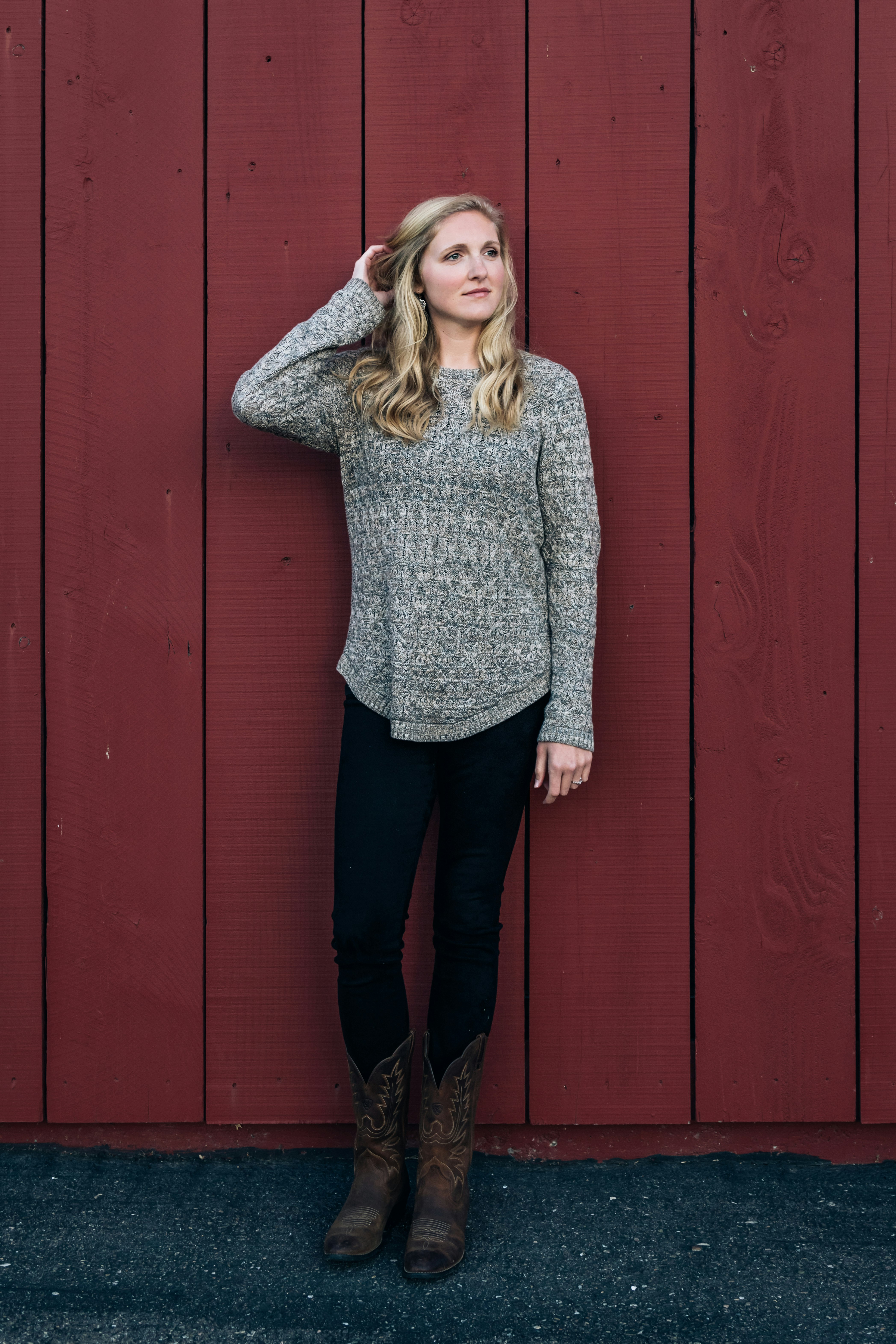 woman in gray sweater and black pants standing beside red wooden wall